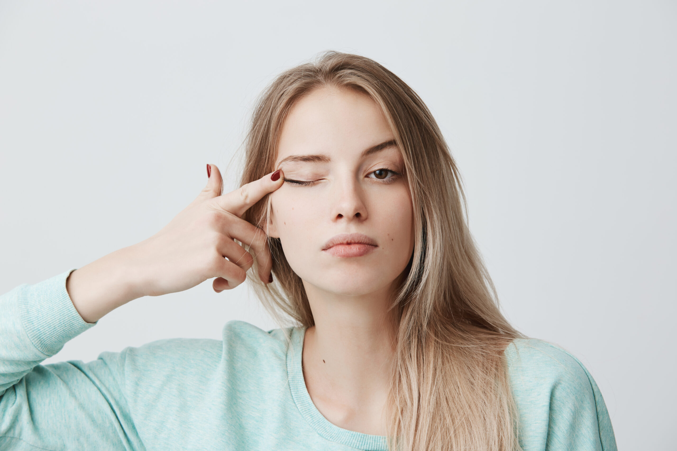 Human face expressions.Thoughtful young beautiful female with blonde dyed straight hair in light blue long-sleeved sweater touching with finger eye, looking at camera with serious and calm expression.