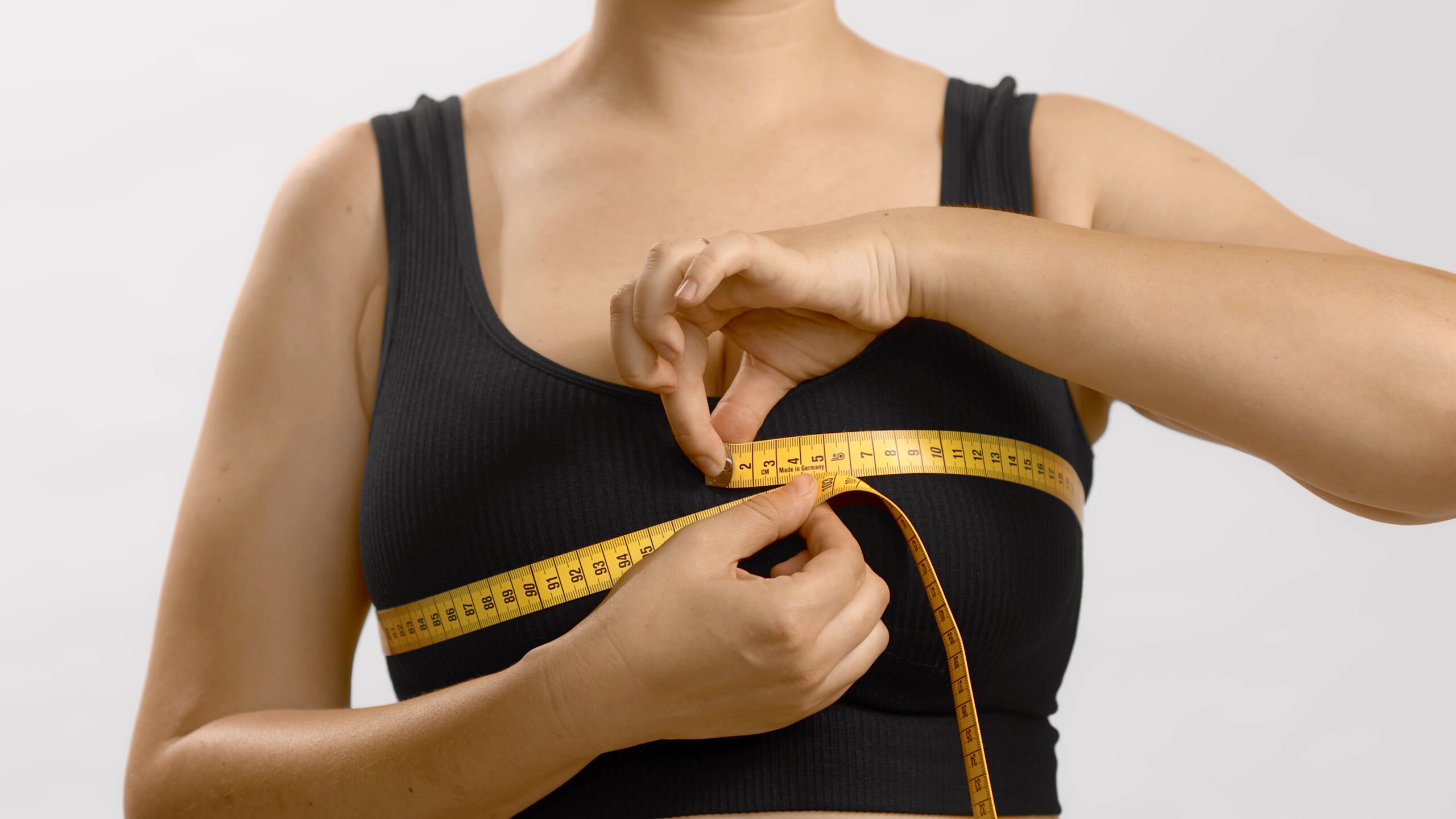 Caucasian young woman in black sport bra measuring her breast with a tape. Studio high quality photo on white background.