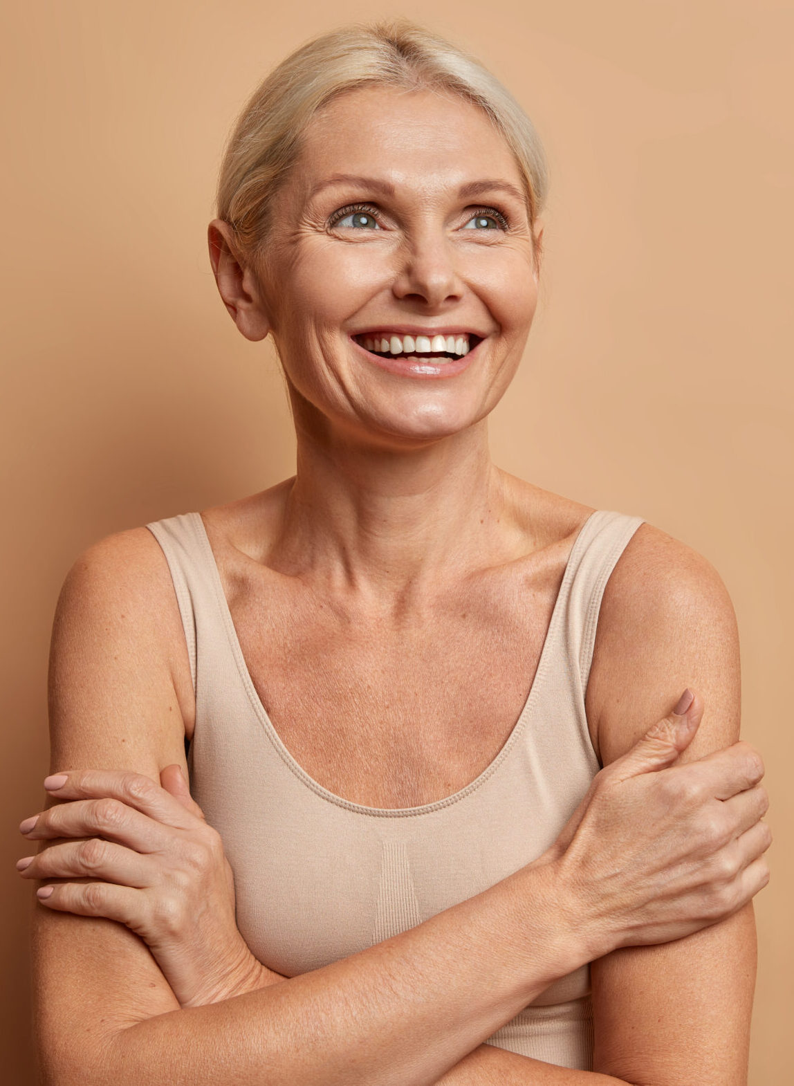 Attractive positive wrinkled fifty years old woman looks gladfully above keeps arms foded has well cared complexion healthy skin white teeth isolated over brown background. Beauty and age concept