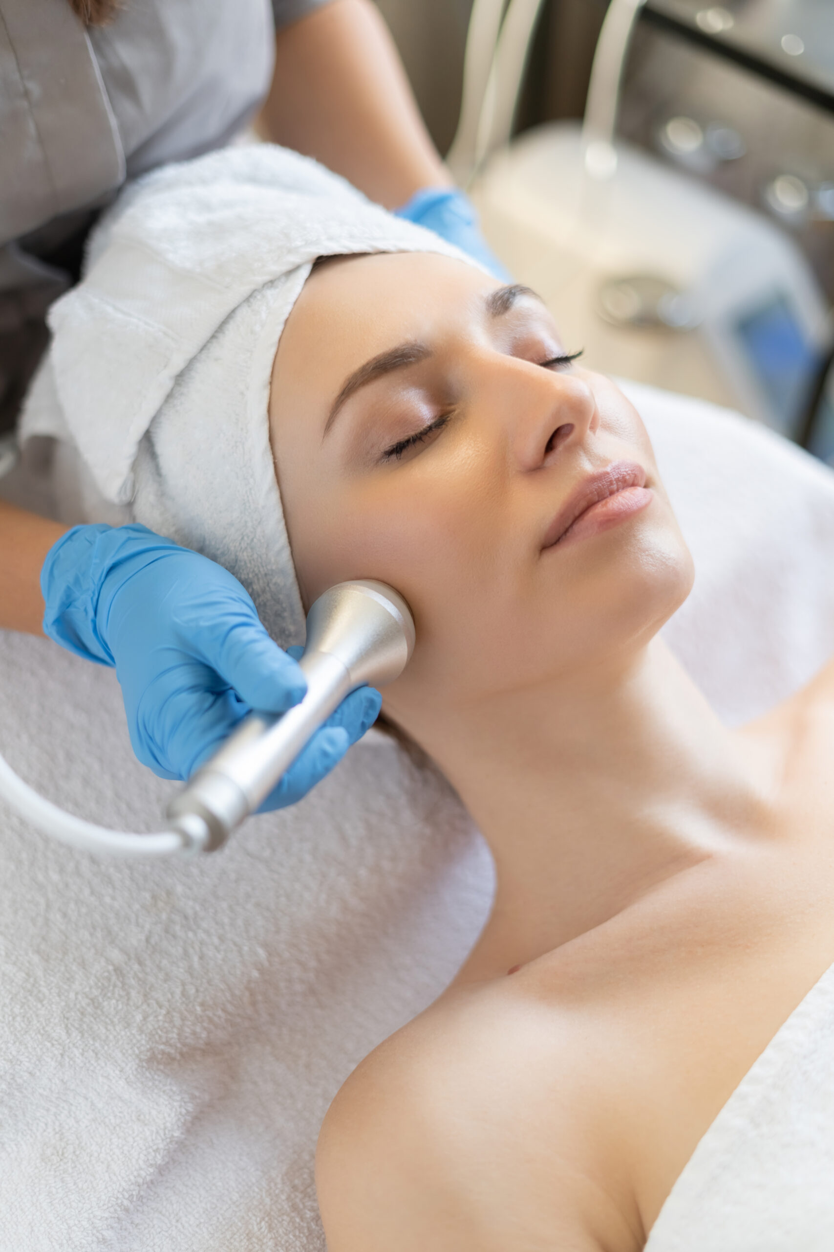 Young woman lying on cosmetologist’s table during rejuvenation procedure. Cosmetologist take care about neck and face skin youthfull and wellness. Hardware face cleaning procedure